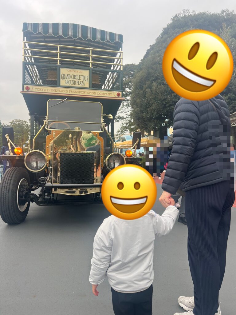 東京ディズニーランドのメインストリートを走る二階建てバス「オムニバス」と、パレードルートで手をつないで見つめる親子の後ろ姿の写真。